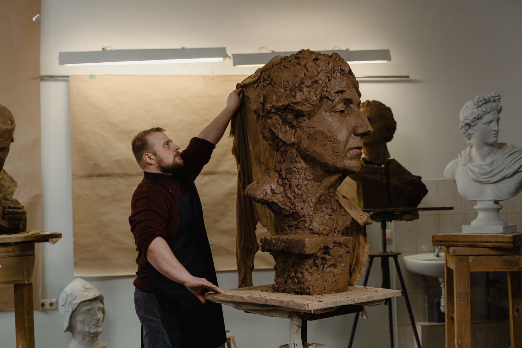 Sculptor working beside a monumental hand-modelled clay portrait in the studio